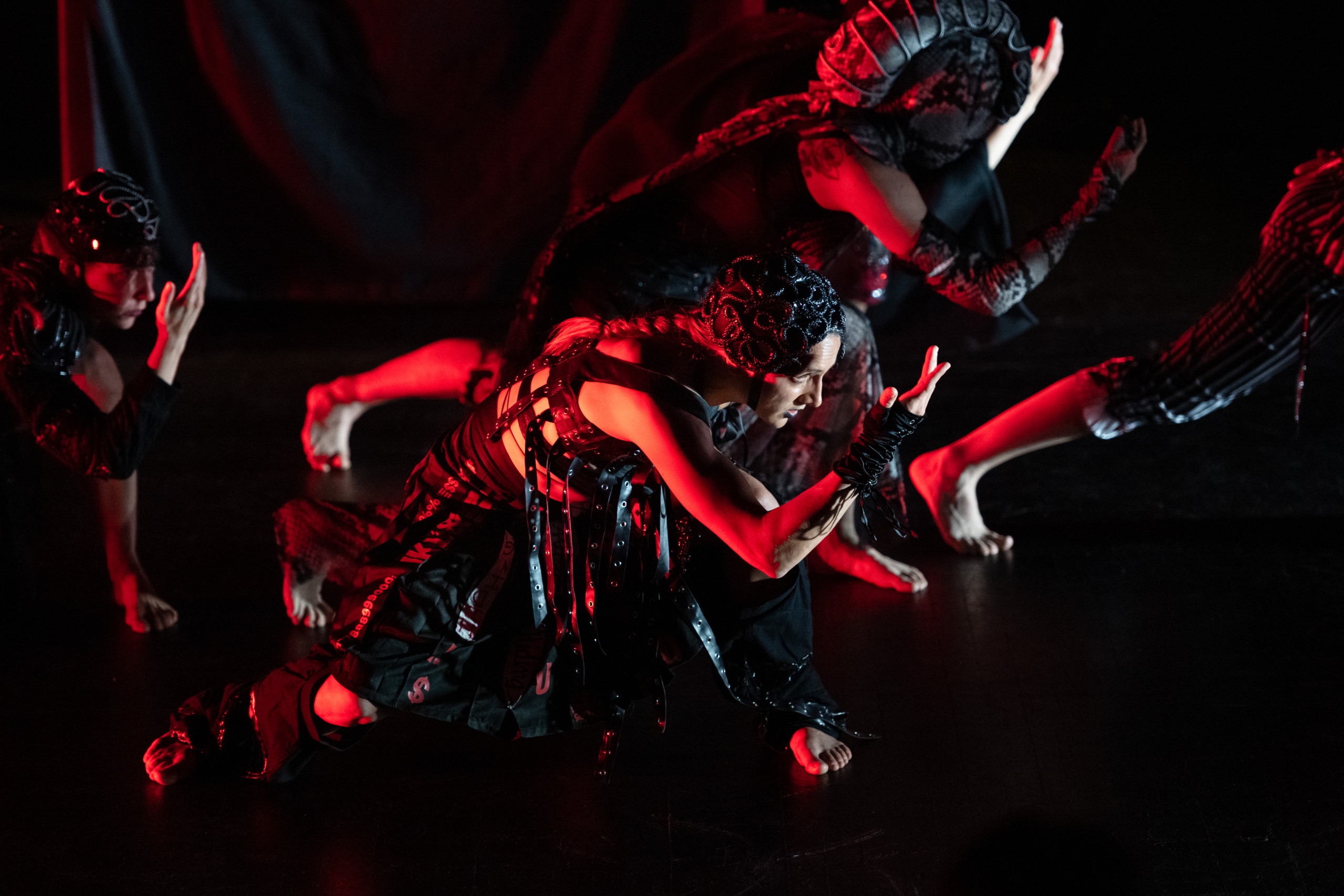 four dancers in warriors/Goth suits lounging to the right, gazing at their right hand, lit by red light on black background.